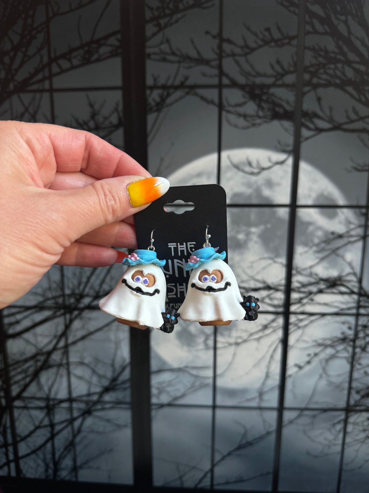 Handmade ghost nugget buddy dangle earrings in a white sheet with a blue brim hat and a small black cat, held against a full moon background