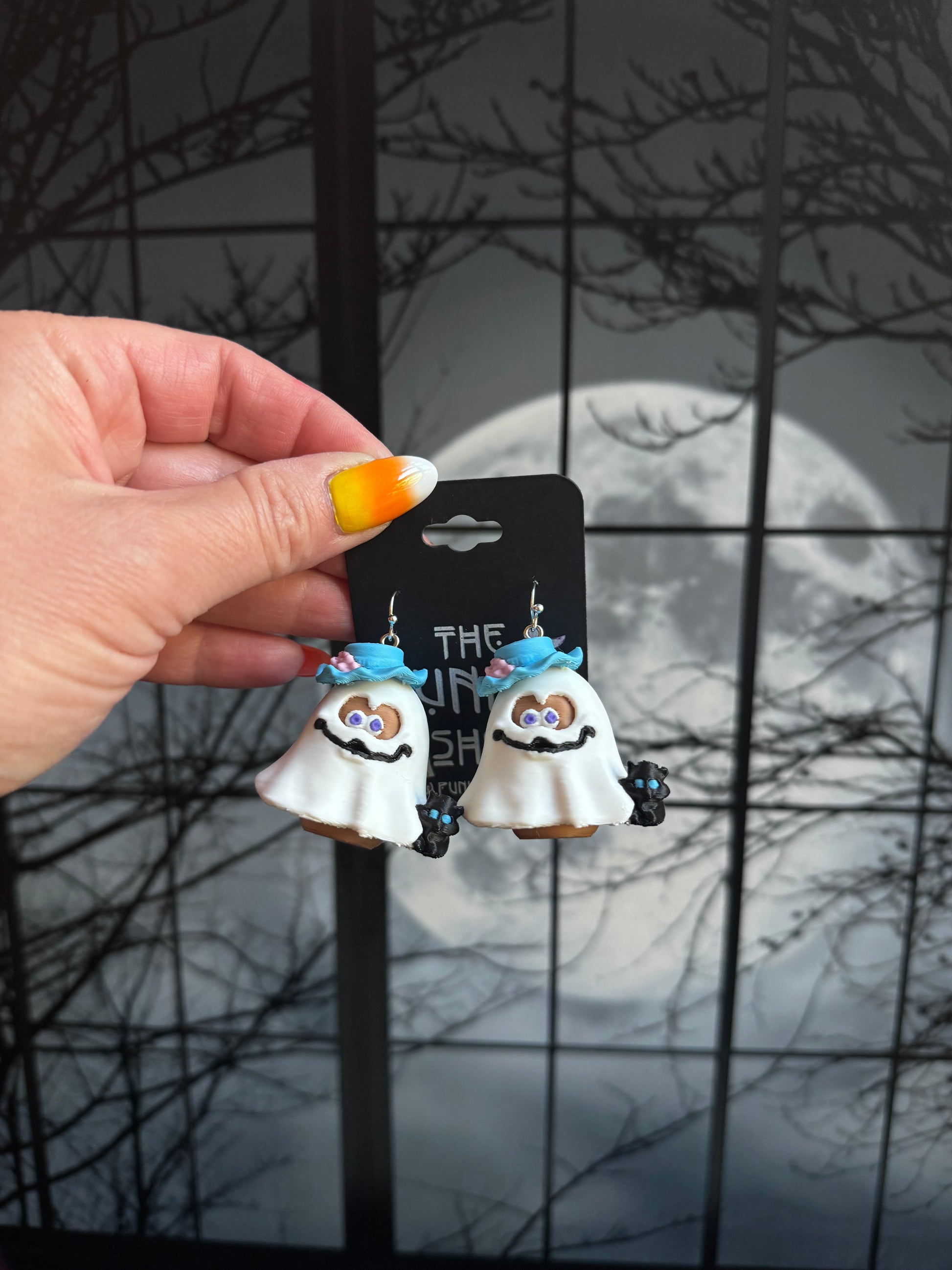 Handmade ghost nugget buddy dangle earrings in a white sheet with a blue brim hat and a small black cat, held against a full moon background
