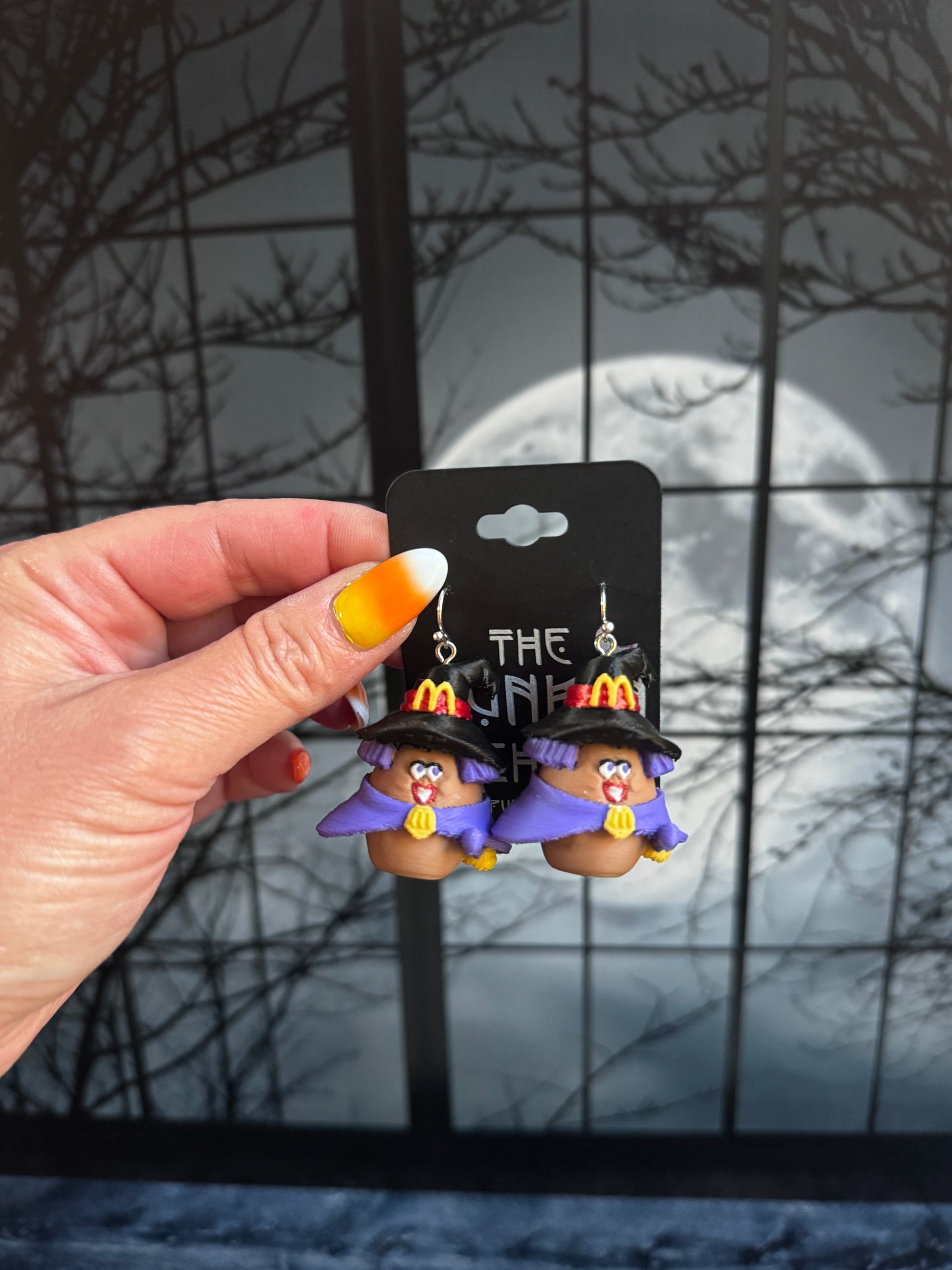 Handmade witch nugget buddy dangle earrings in a purple pointed hat and purple cloak, held against a full moon background